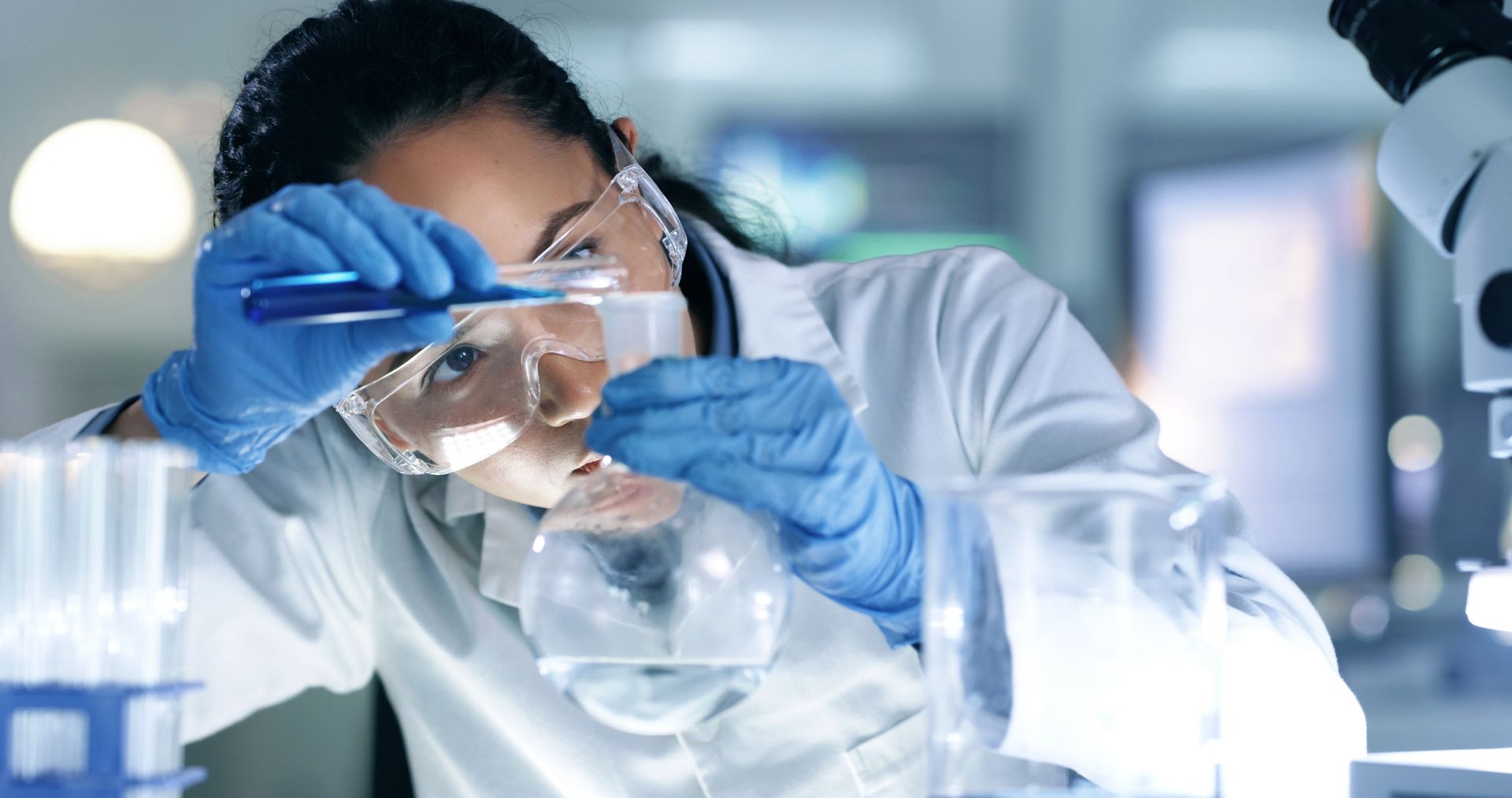Scientist in lab coat, safety glasses, and blue gloves pouring liquid from a vial into a flask.