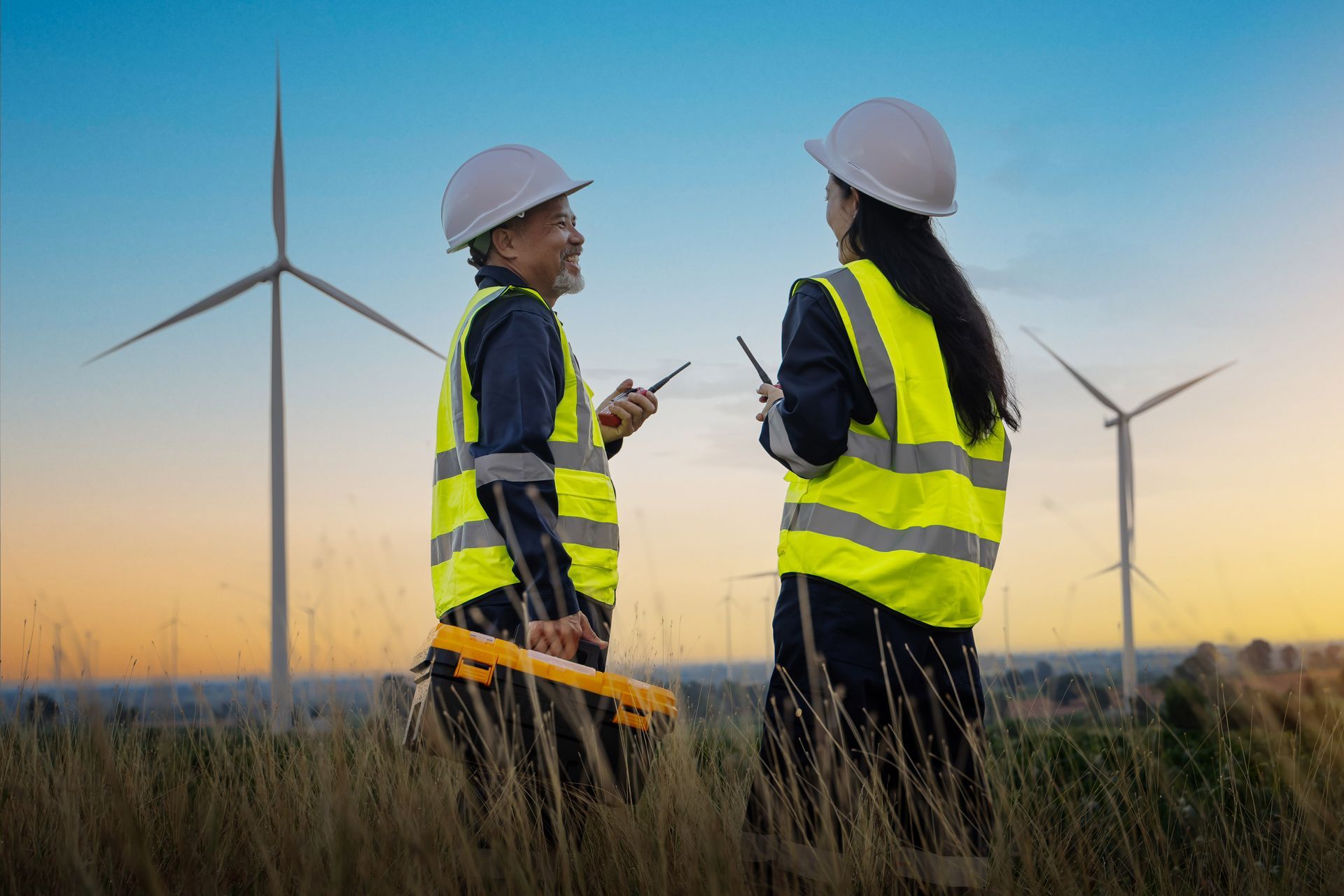 Two people in safety vests and hard hats converse near wind turbines at sunset.