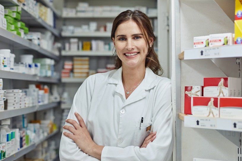 Pharmacist in a white coat, smiling, leaning in a pharmacy aisle surrounded by medications.