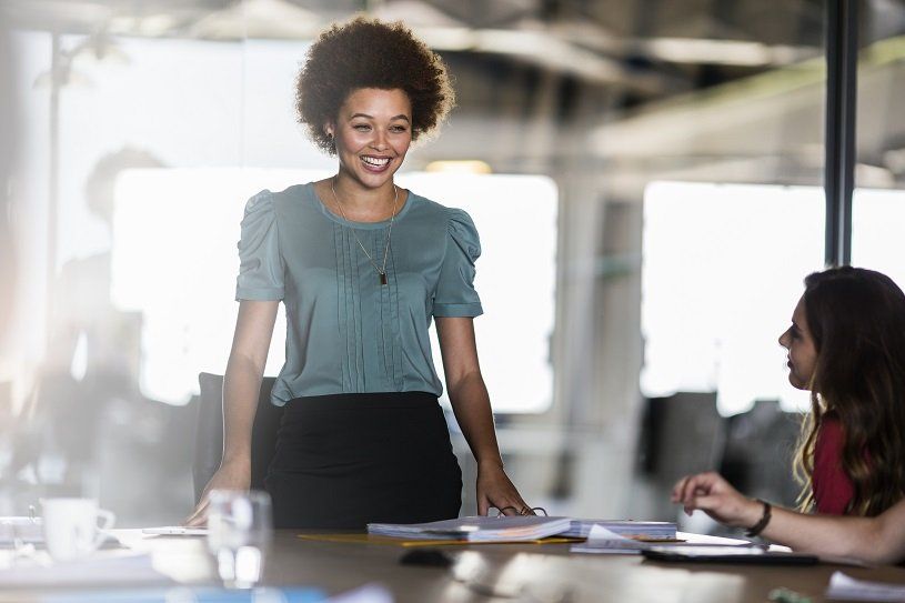 Woman smiling, standing over a table in an office, speaking to another woman seated across.