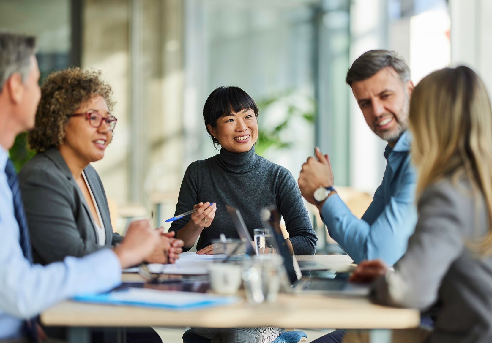 Business team in meeting, smiling, discussing at a table with laptops and documents. Natural light setting.