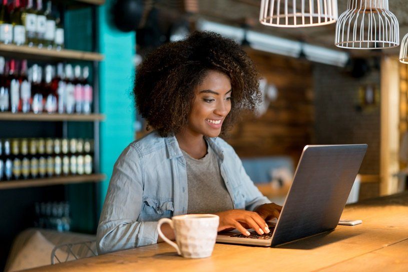 Woman working on laptop at a cafe, smiling, with coffee, near shelves of bottles.