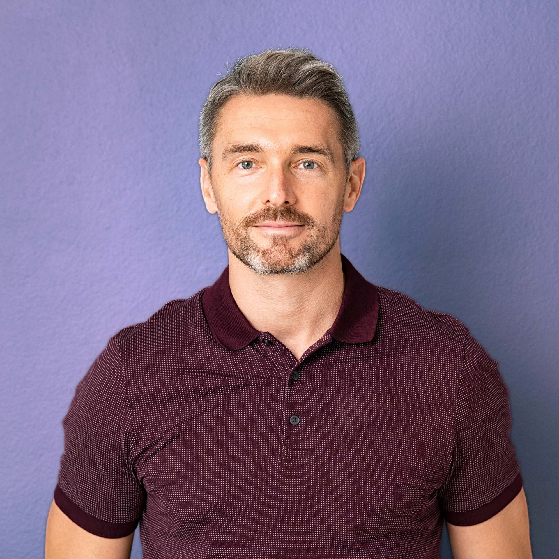 Man with short gray hair and stubble, wearing a burgundy polo shirt, smiles against a purple background.