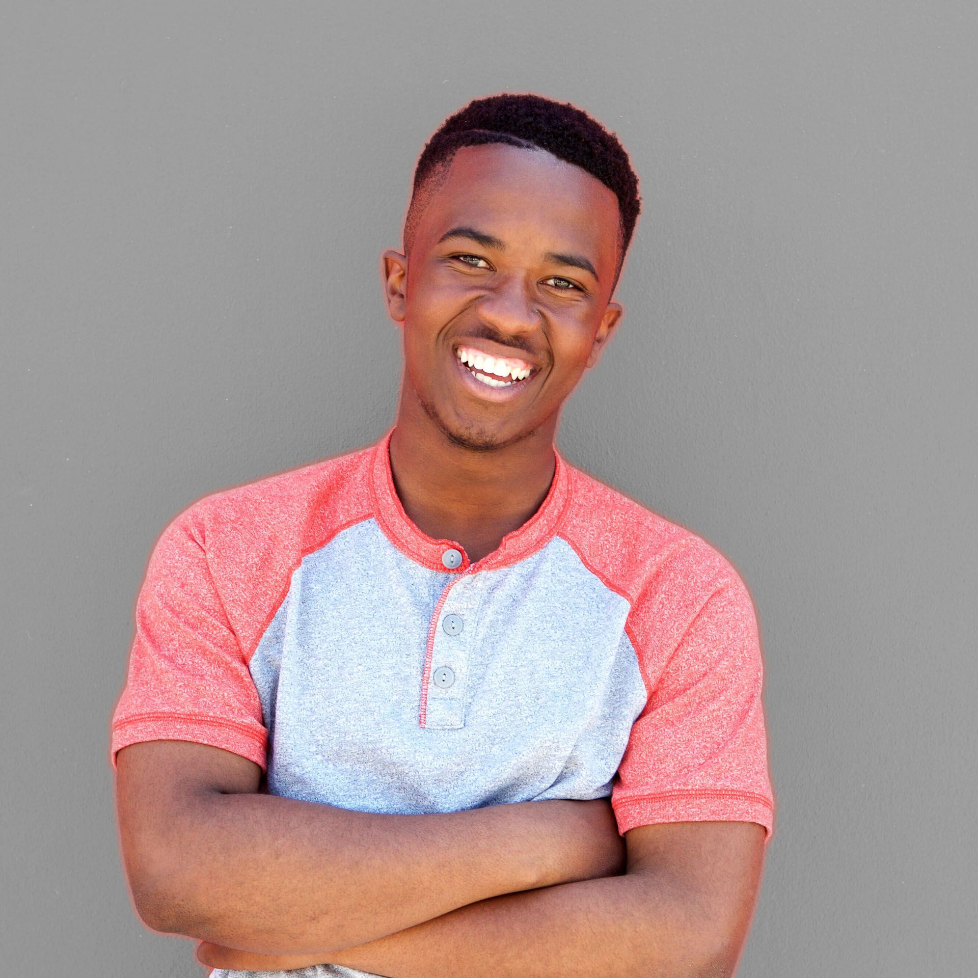 Man with arms crossed smiling, wearing a red and gray shirt, against a gray background.