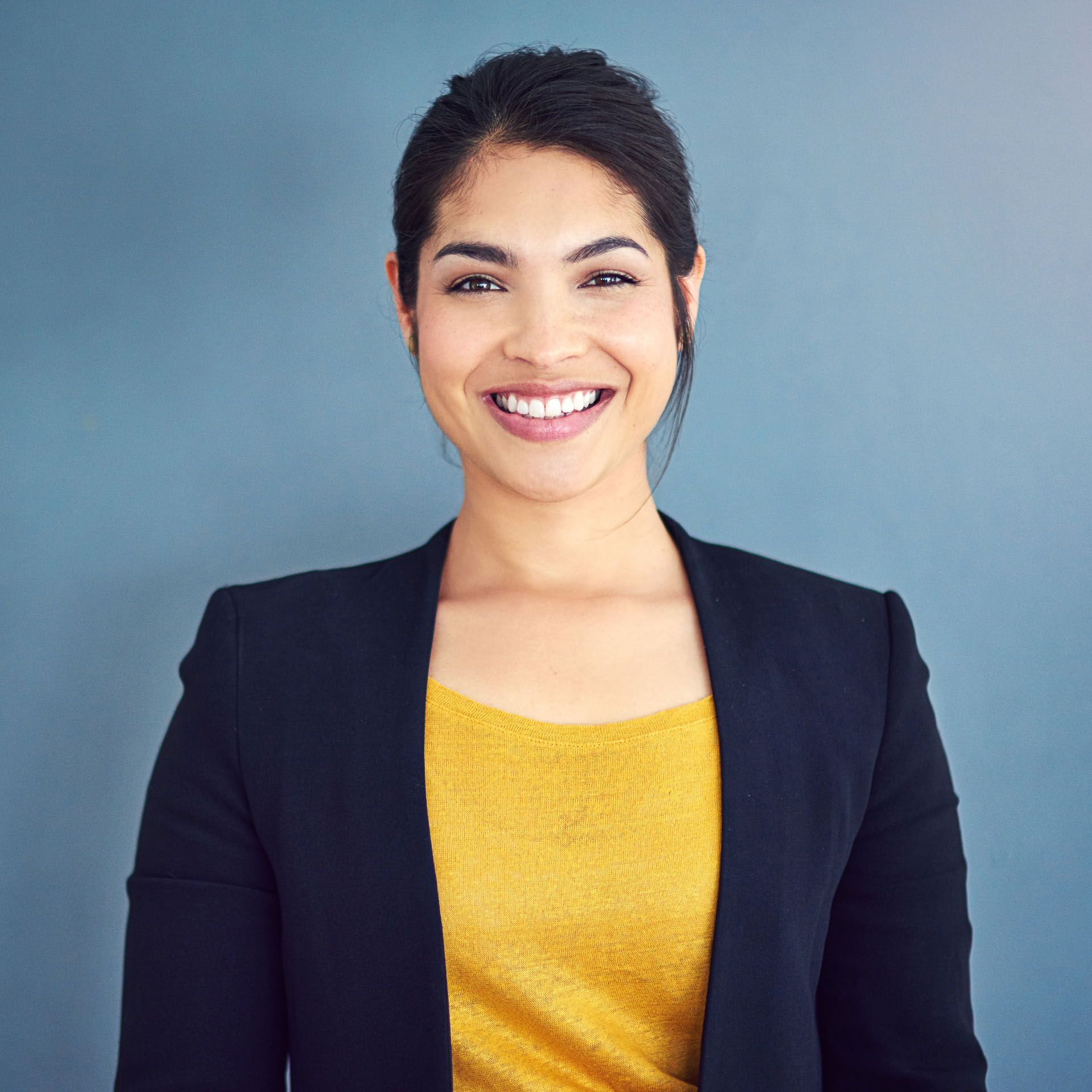 Woman smiling, wearing a yellow top and black blazer against a blue background.