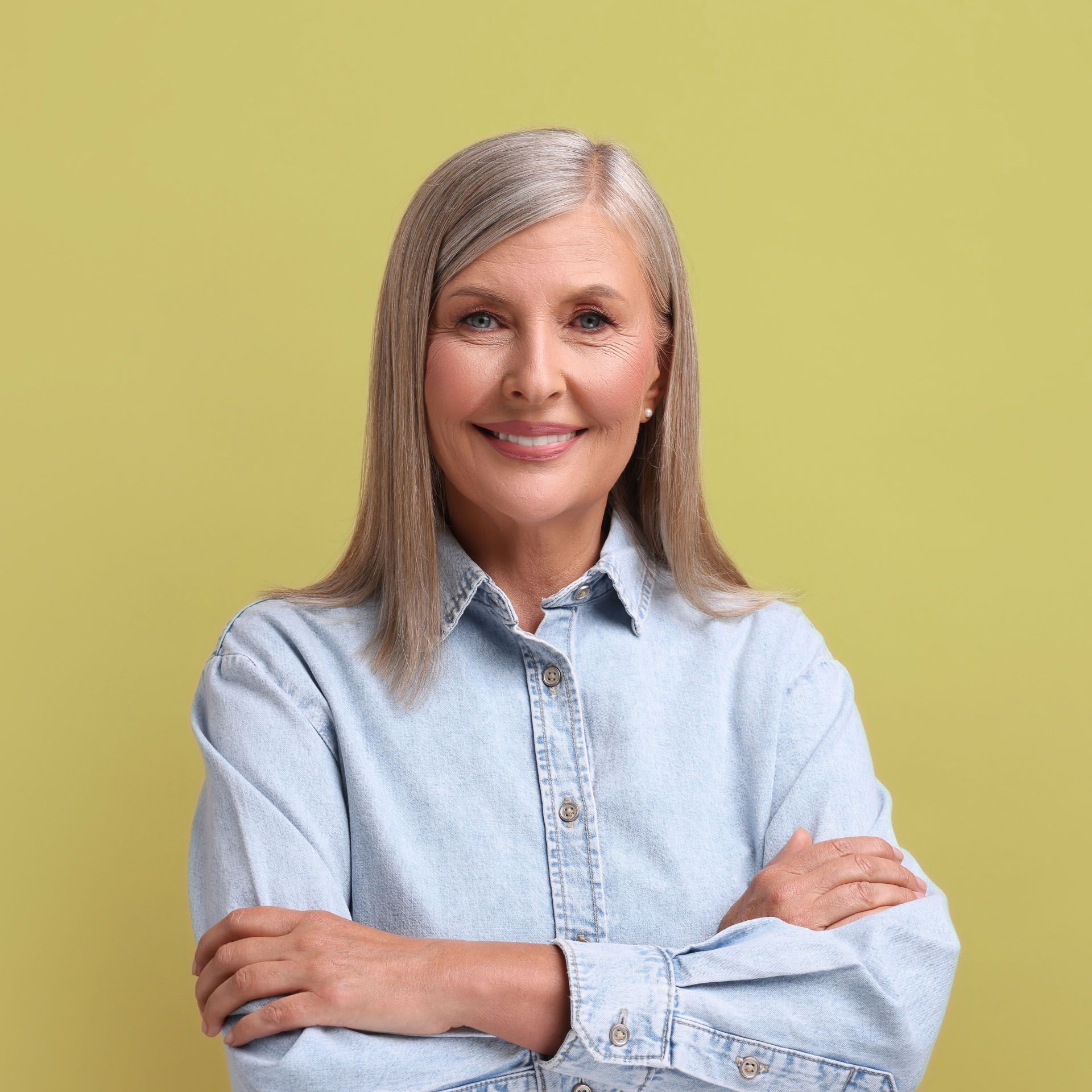 Woman with silver hair, smiling with arms crossed, wearing a denim shirt, against a yellow background.