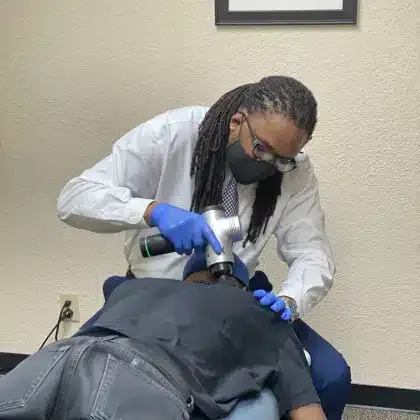 Clinician in white coat and blue gloves using a handheld treatment device on a patient lying face down on a table