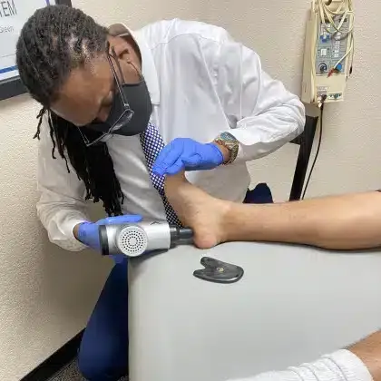 Healthcare worker in white coat and blue gloves using handheld device on a patient’s leg in a clinic room