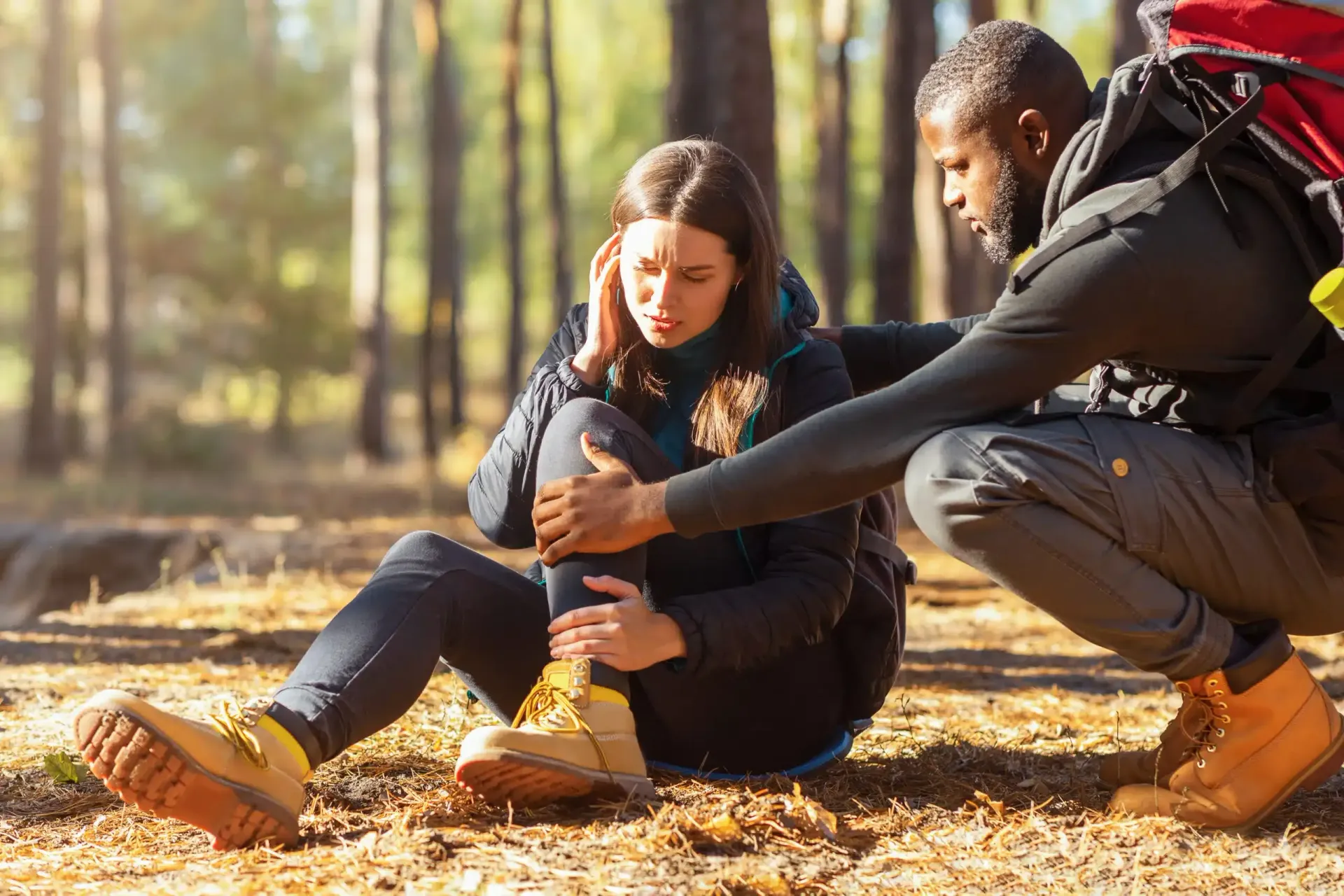 Two hikers sitting on a forest trail, one tying the other’s boot in warm sunlight.