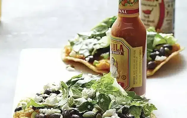 Tostadas topped with beans, lettuce, and cheese, with a hot sauce bottle in the foreground.