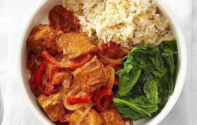 Bowl of rice with red saucy meat, red peppers, and wilted greens
