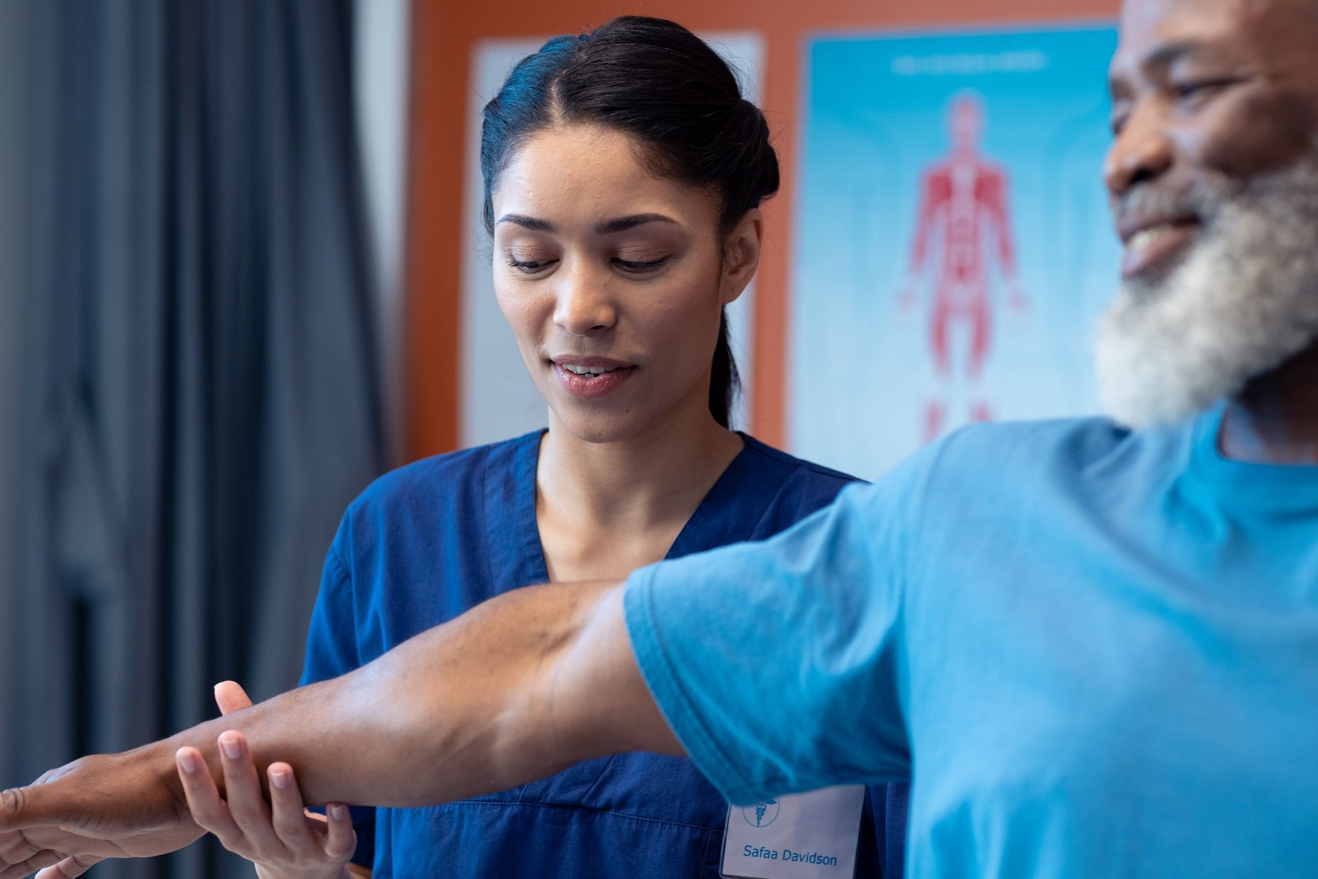 Physical therapist in blue scrubs examining a patient’s arm in a clinic with anatomy poster in the background