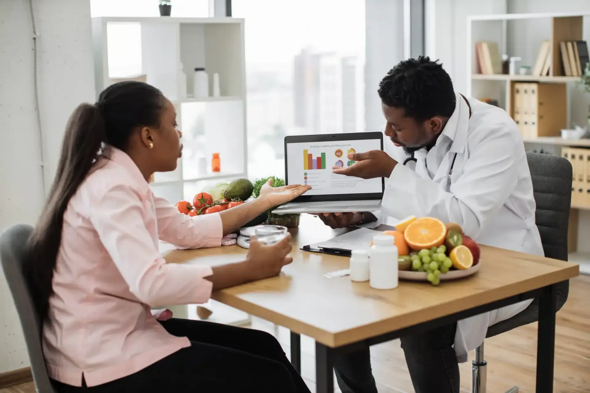Two people discussing nutrition at a table with a laptop and fruit in a bright office-like room