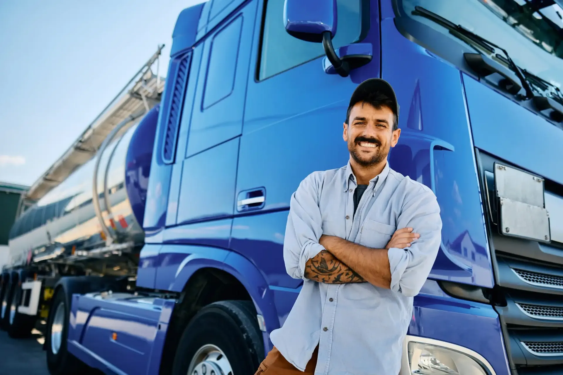 Smiling man in light shirt standing with arms crossed beside a blue cargo truck