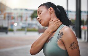 Woman in a green tank top clutching her neck, appearing to feel pain outdoors.