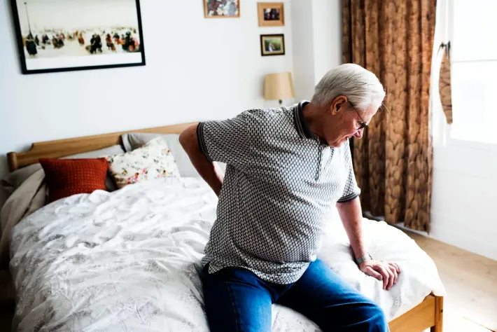 Person sitting on the edge of a bed in a bright bedroom, looking toward the window.