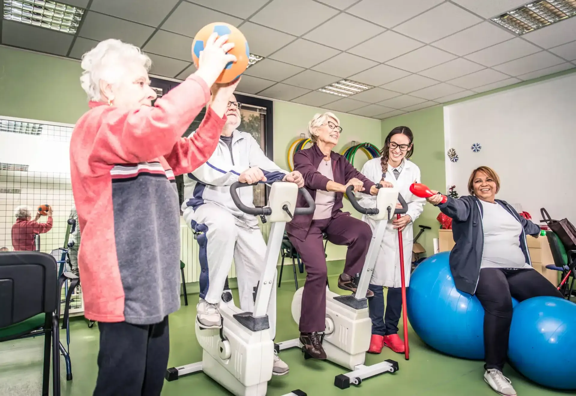 Seniors exercise together in a therapy room using walkers and blue stability balls.