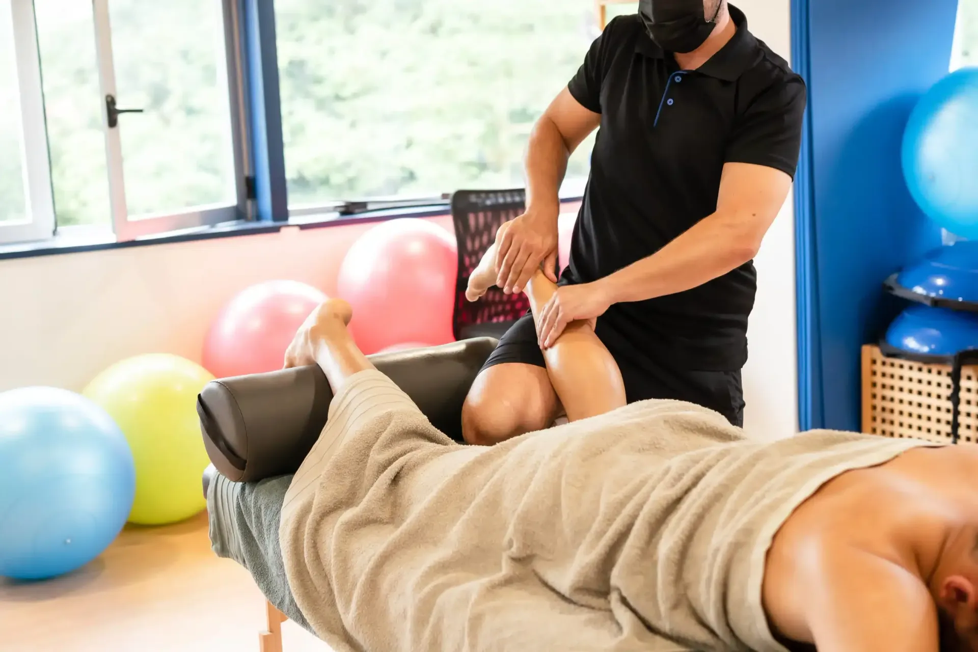Massage therapist working on a client’s leg in a bright therapy room with exercise balls.