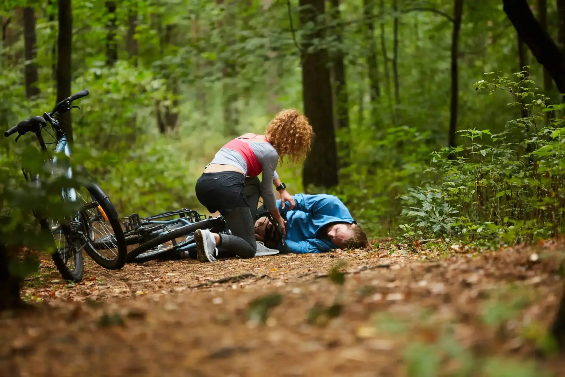 Two cyclists kneel beside a fallen bike on a forest trail, helping an injured rider in blue.