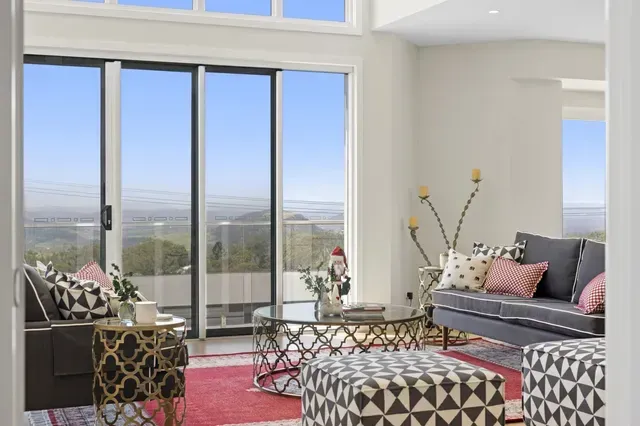 Bright modern living room with large windows, geometric chairs, gray sofa, pink rug, and ocean view