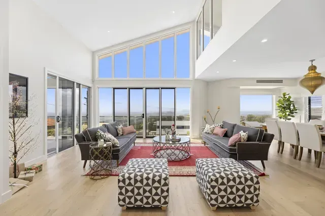 Bright modern living room with floor-to-ceiling windows, gray sofas, red rug, and ocean view