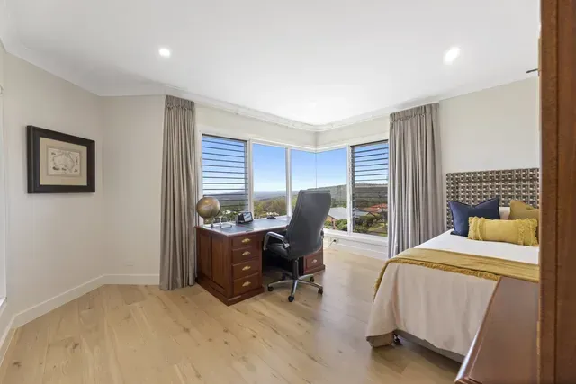 Bright bedroom with desk by corner windows, bed on the right, and light wood floors.