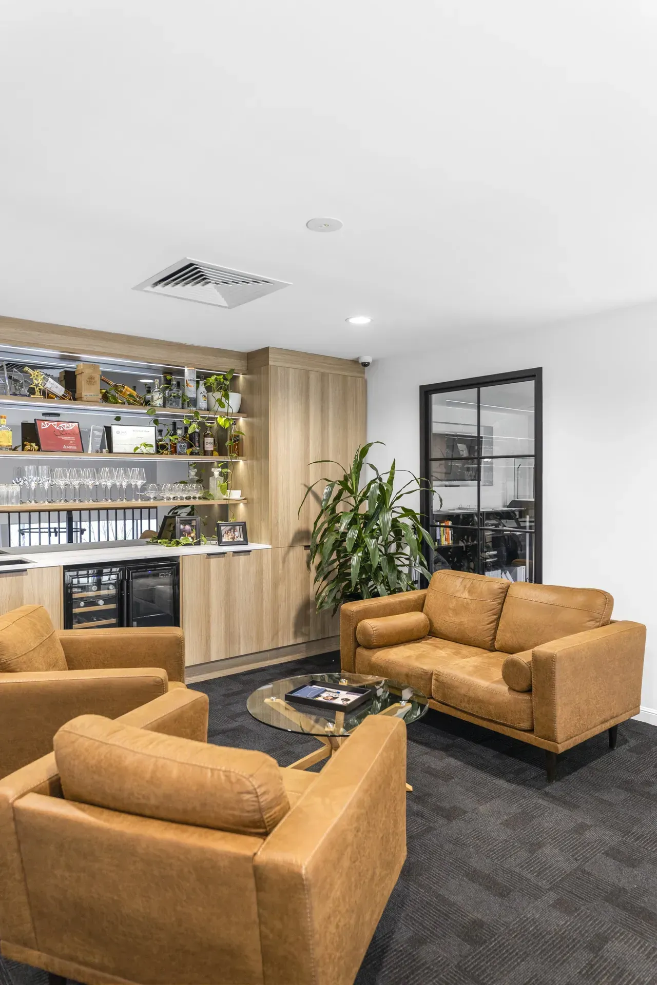 Modern office lounge with tan chairs, dark carpet, wooden shelving, and a potted plant.