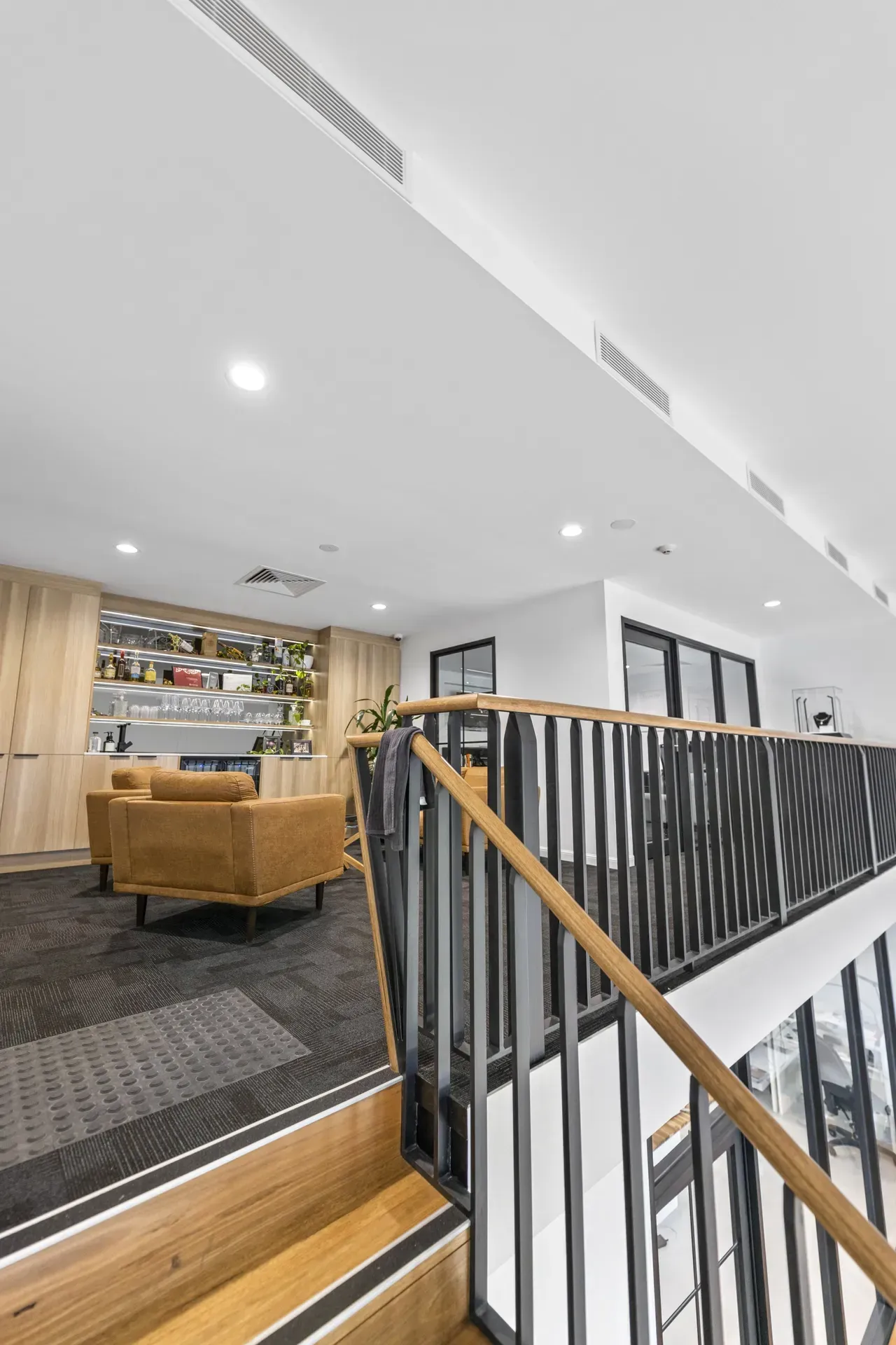 Modern lounge with wooden staircase, black railing, tan sofa, and open seating area