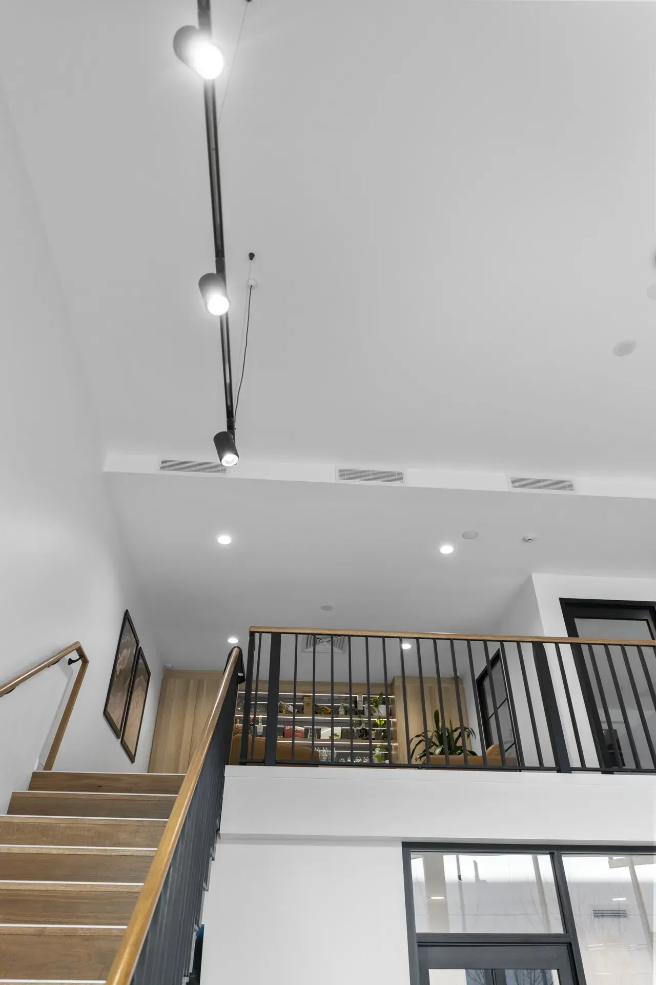 Modern interior staircase with wooden steps, black railing, and ceiling track lights beneath a mezzanine.
