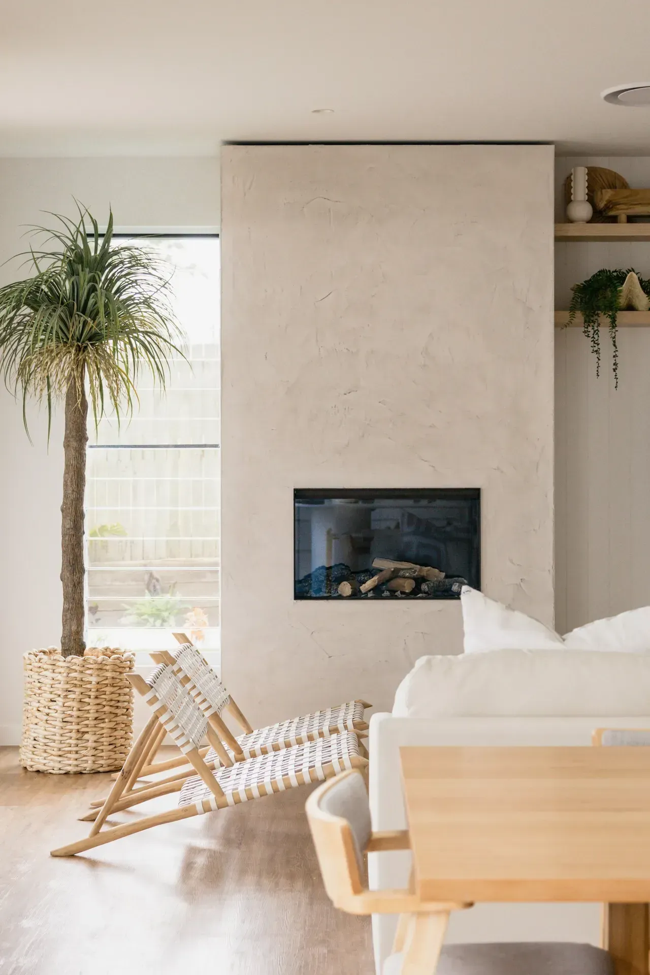 Cozy living room with a fireplace, tall plant, woven chair, and light wood furniture
