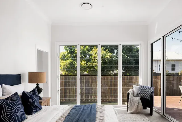 Bright bedroom with large windows, bed, nightstand, chair, and sliding glass door to a deck.