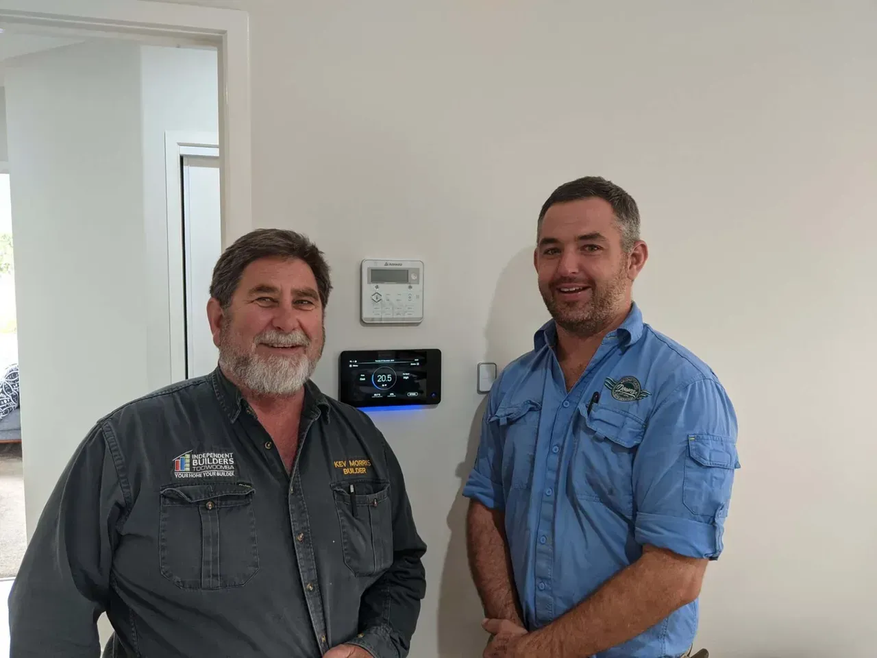 Two men in work shirts standing indoors beside a wall-mounted thermostat and control panel
