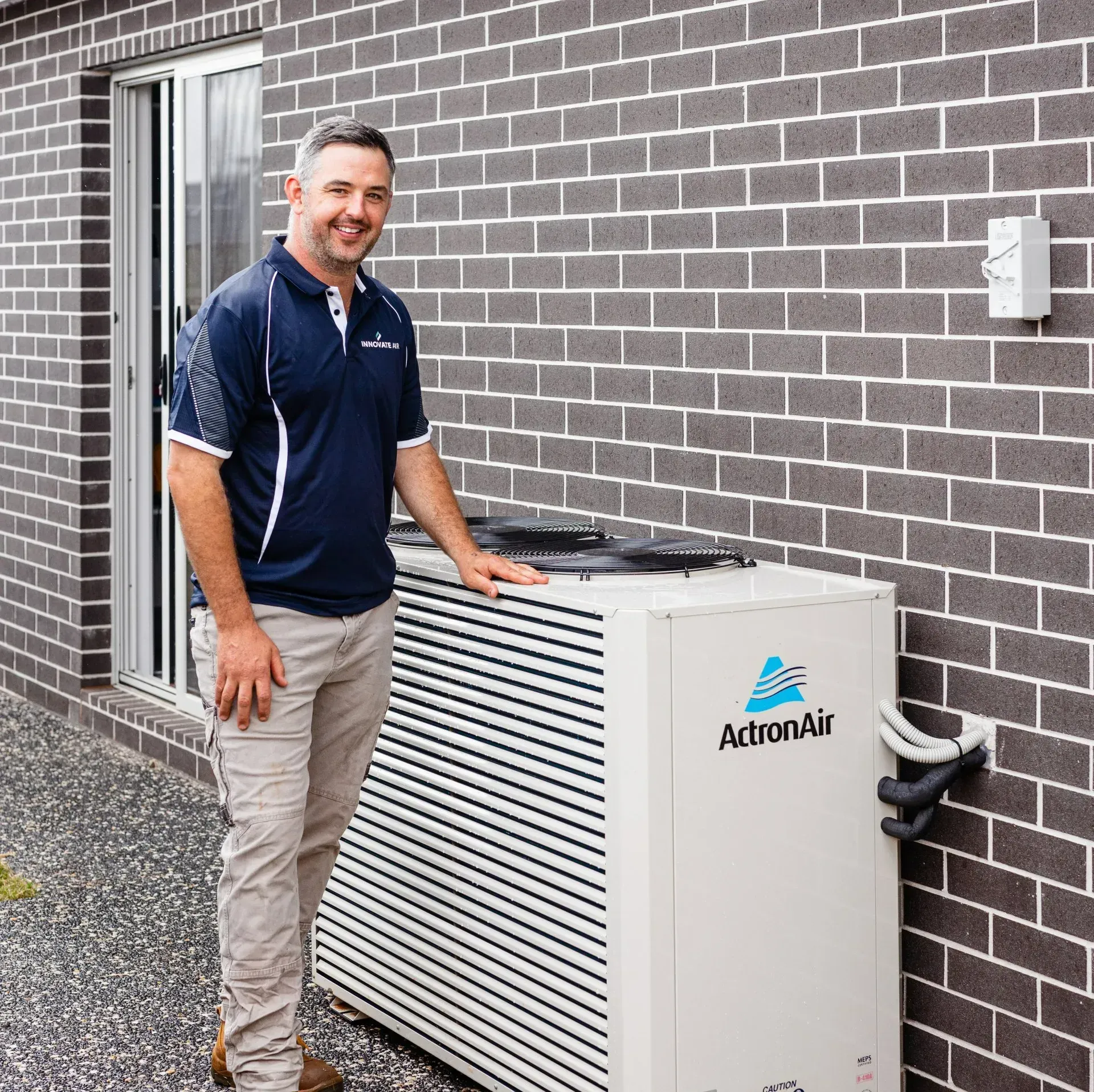 Technician standing beside a white ArtronAir outdoor unit against a brick wall