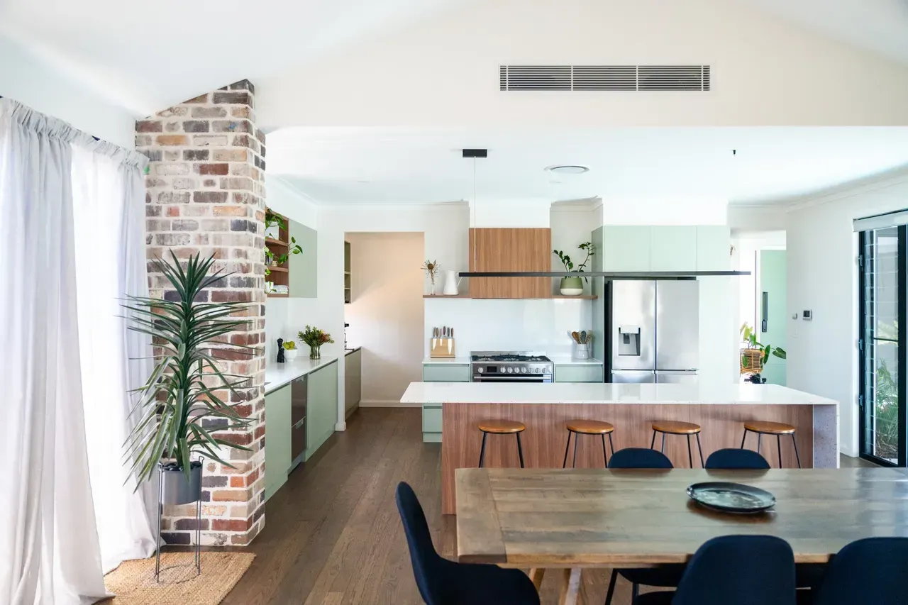 Bright modern dining and kitchen area with wood table, brick column, white cabinets, and large windows