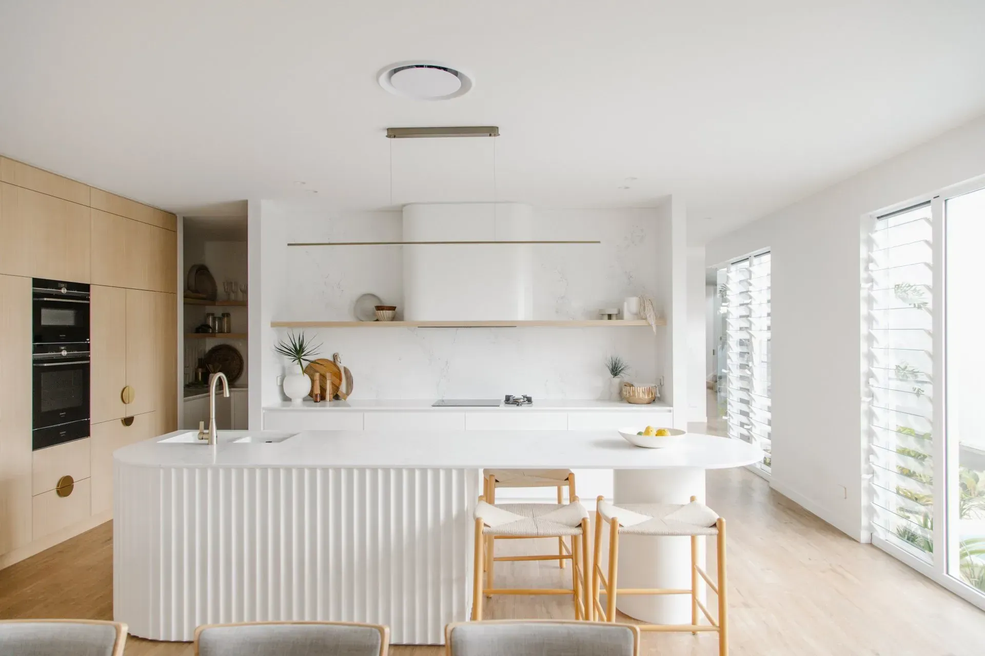Bright modern kitchen with white cabinets, island, wooden stools, and sunlight from tall windows