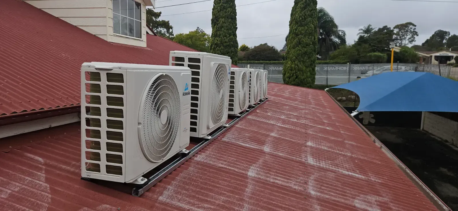 Outdoor air conditioner units lined along a red corrugated roof beside a blue canopy