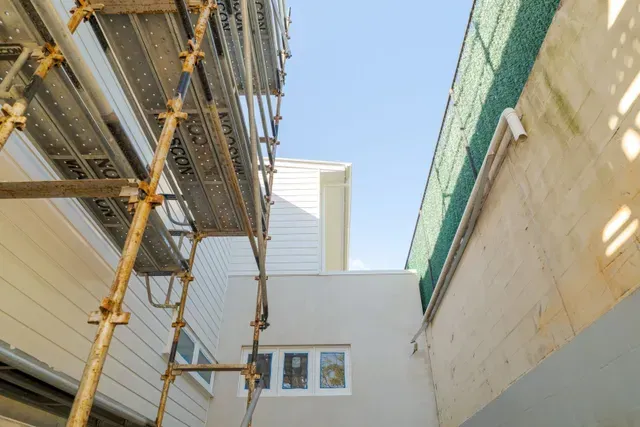 Scaffolding in a narrow courtyard between tall buildings, with a white wall and blue sky overhead