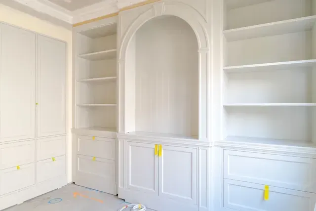 White built-in cabinets and shelves in a bright room, with masking tape on the doors and floor