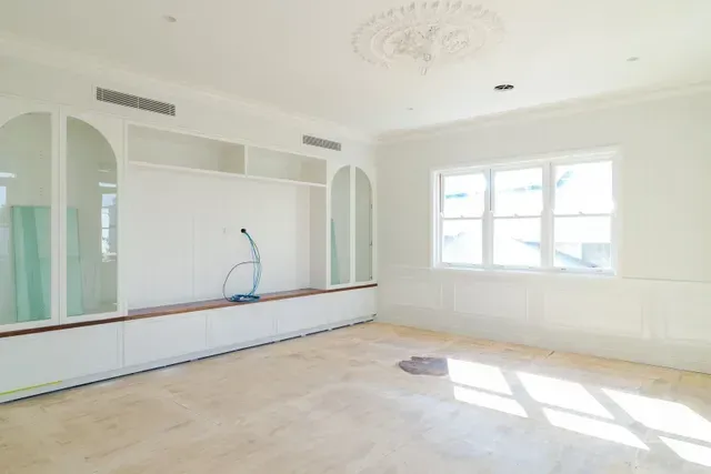 Bright empty room with white walls, built-in shelving, large window, and exposed floor under renovation