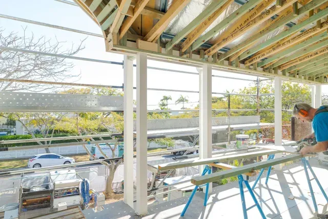 Construction site with exposed roof framing and workers on scaffolding, bright daylight outside