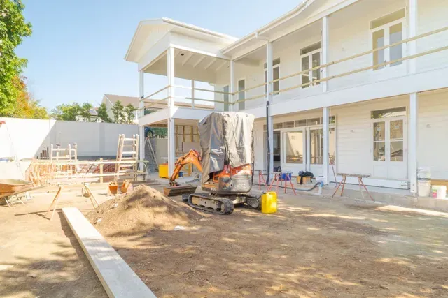 Construction site with a two-story white house, workers, and dirt piles in the yard