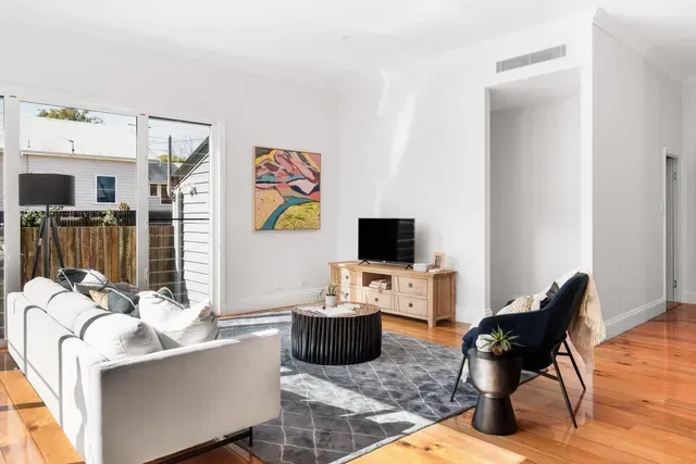 Bright living room with white sofa, black chairs, TV on wooden stand, and patio doors to a sunny deck.