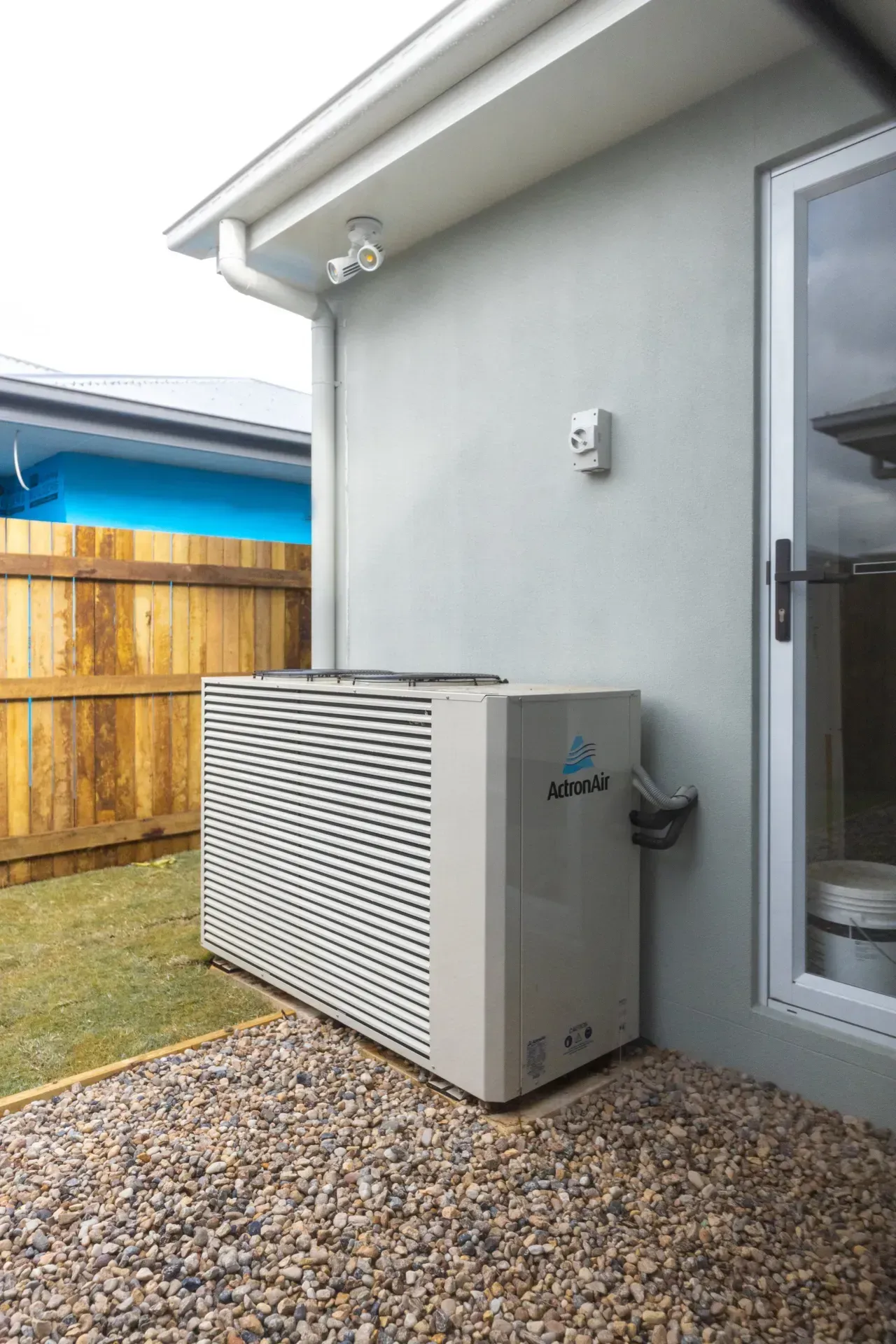 Outdoor air-conditioning unit beside a gray house wall under an eave, next to a glass door