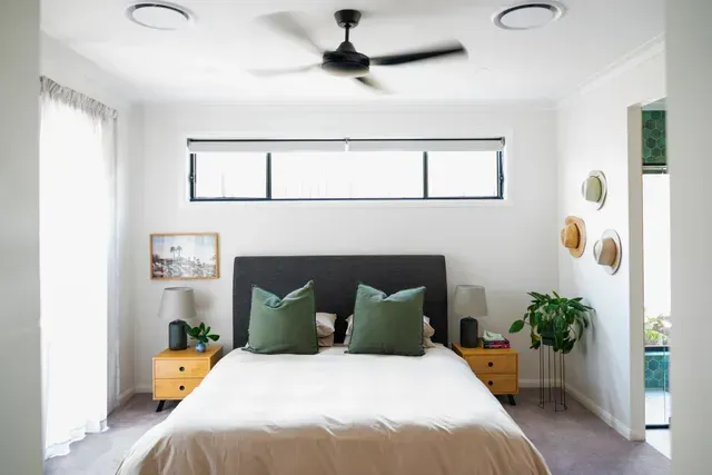 Bright minimalist bedroom with a bed, gray headboard, green pillows, and wood nightstands under a ceiling fan.