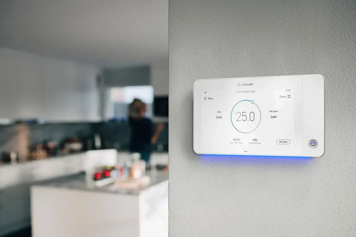 White smart thermostat on a gray wall, glowing blue, with a blurred kitchen in the background