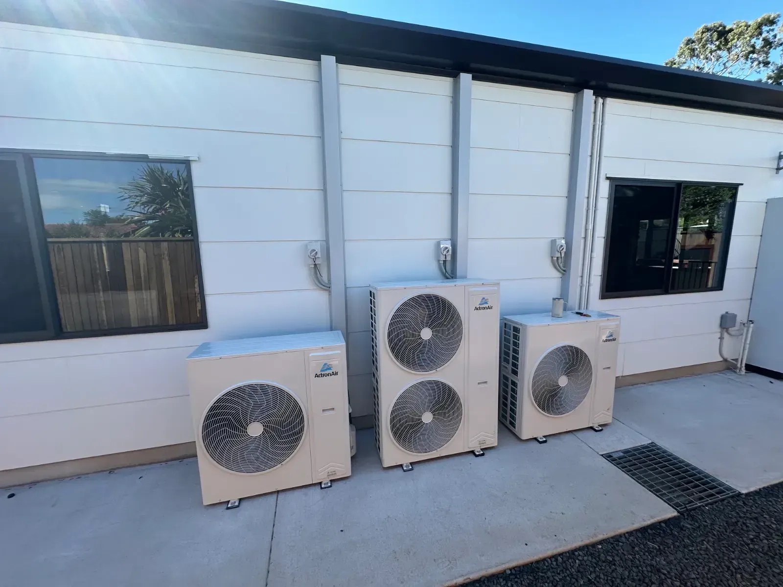 Three outdoor HVAC condenser units against a white building wall beside windows and a doorway.