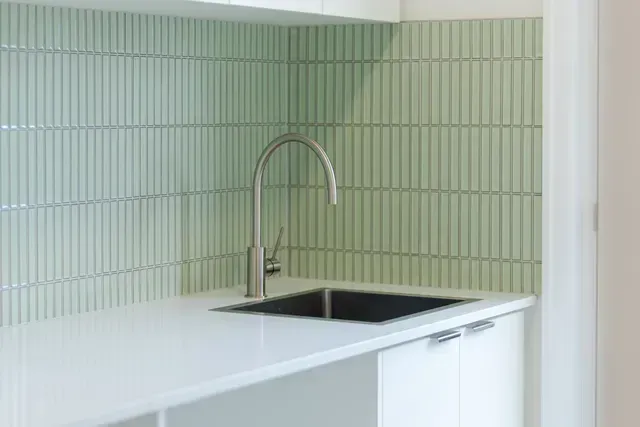 Minimalist kitchen corner with pale green tile backsplash, white countertop, black sink, and chrome faucet