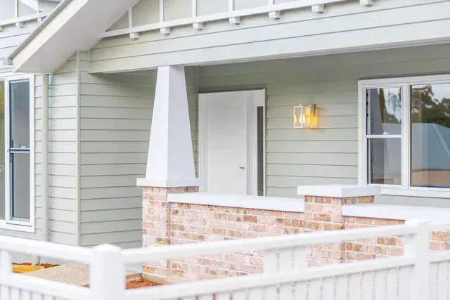 Covered front porch with pale green siding, white railing, and brick half walls beside a lit wall sconce.