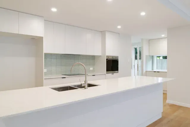 Bright modern white kitchen with island, sink, built-in oven, and recessed ceiling lights
