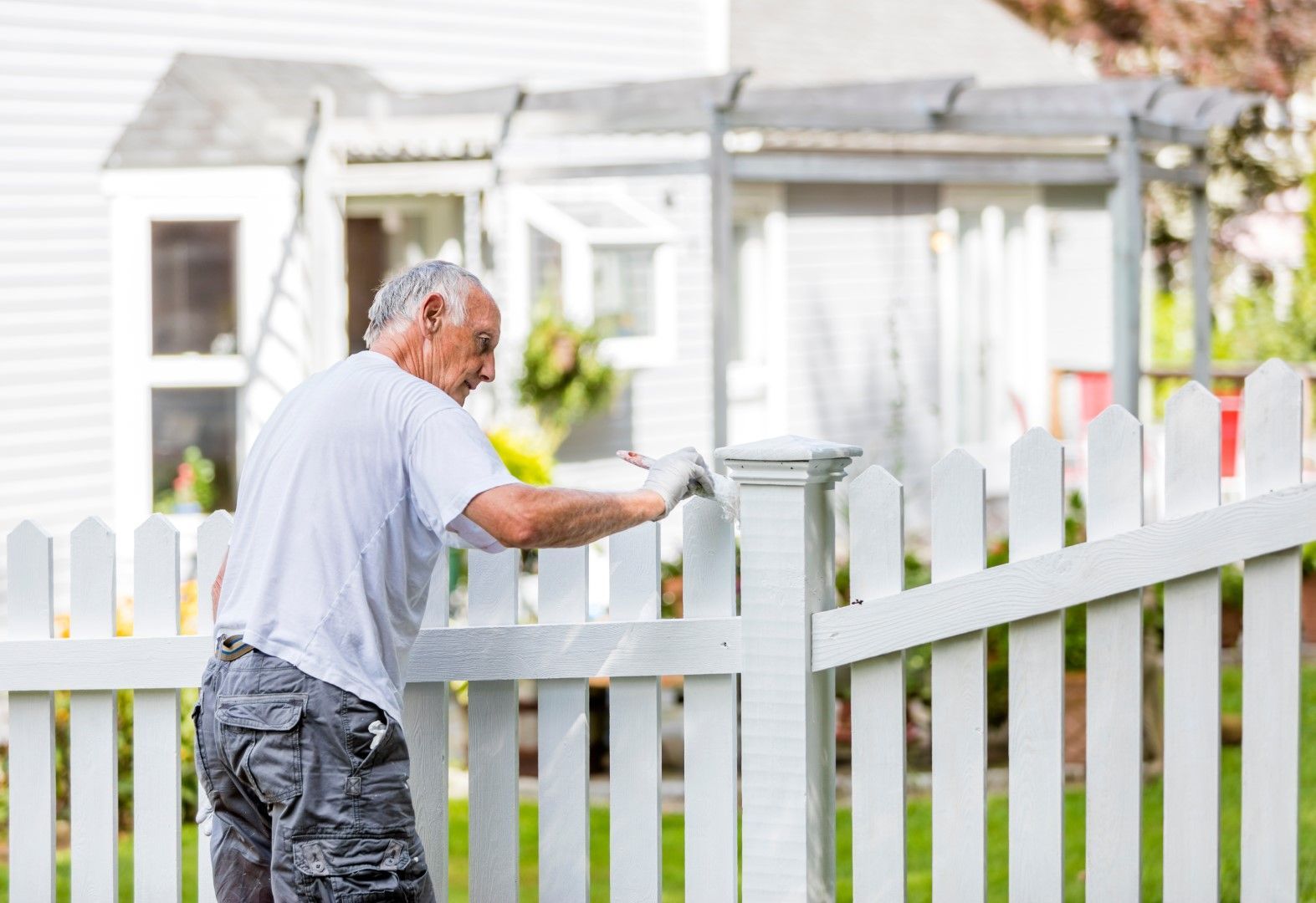 Older man painting a white picket fence in front of a house.